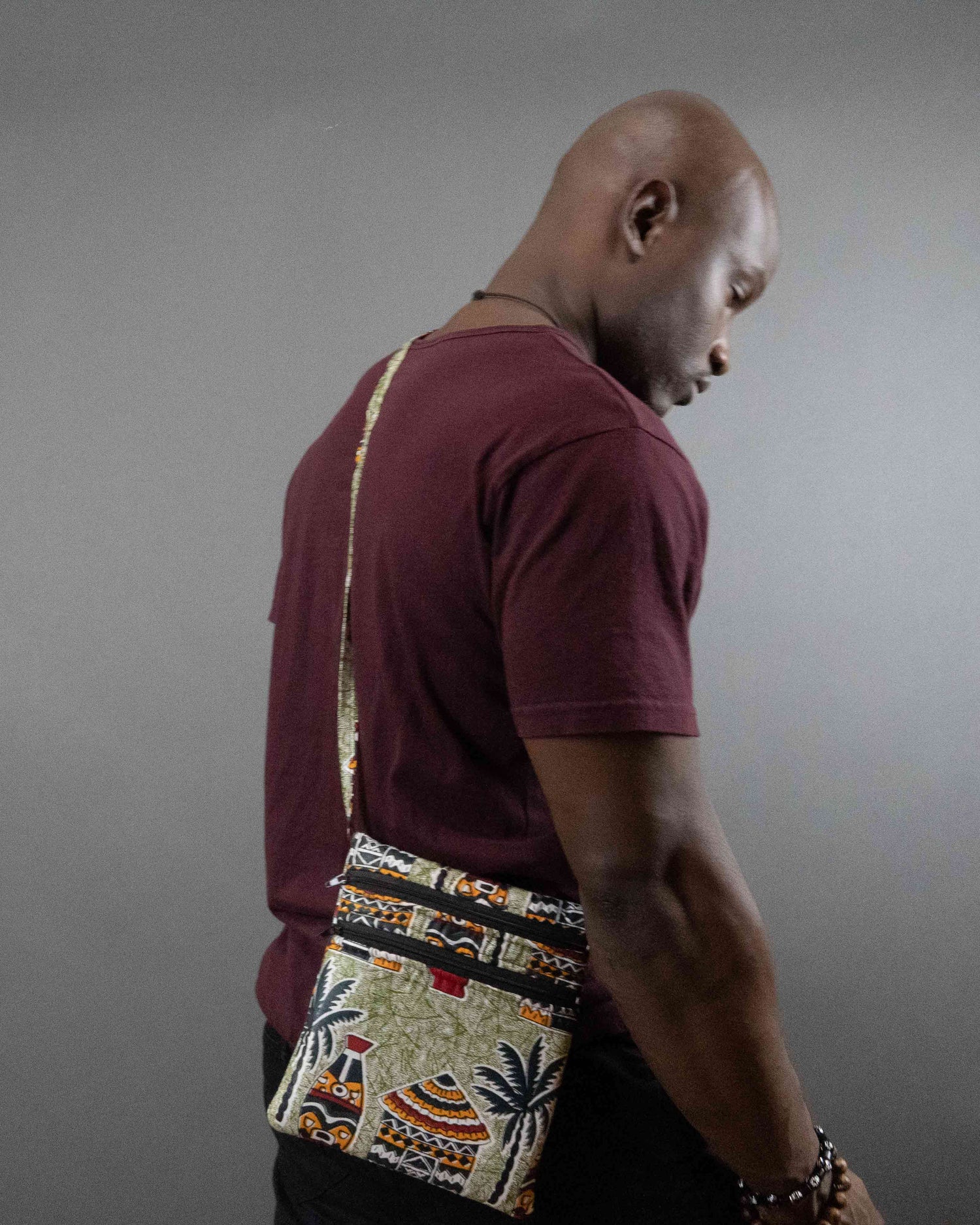 This is a photo of a dark skinned African model wearing a Juste Design crossbody bag-Home. The model is muscular, and has a maroon shirt on; his head is tilted downward like that of a man in deep deep thoughts